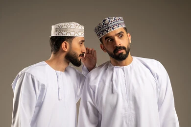 مشاركة الاسرار مع الأصدقاء، التحدث بصوت منخفض، بورتريه لرجلين عربيين خليجيين عمانيين يرتديان الدشداشة و الكمة و يقوم أحدهما بالهمس في اذن صديقه, خلفية رمادية