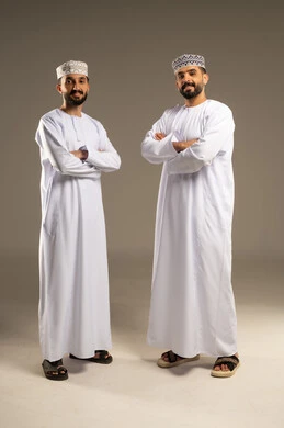الوقوف باستقامة و ايدي متقاطعة، مفهوم الرجولة و الثقة بالنفس,  بورتريه لرجلين عربيين خليجيين عمانيين يرتديان الدشداشة و الكمة ينظران الى الكاميرا بايماءات السعادة و السرور, خلفية رمادية