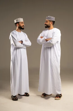 Standing with crossed arms, a portrait of two Arab Gulf Omani men wearing dishdasha and kumma standing side by side, gestures of happiness and joy, gray background.
