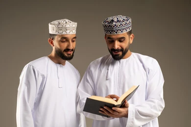 Developing reading skills, a portrait of two Arab Gulf Omani men wearing dishdasha and kumma, one holding a book and reading it, with a gray background.