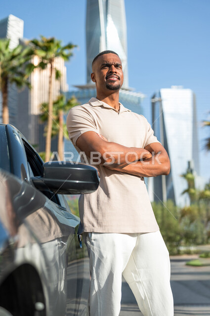 شاب ذو بشرة سمراء عربي خليجي سعودي يقف بجانب سيارته في أحد شوارع الرياض ، الاستمتاع و الإنبهار بجمال المدينة ، قضاء وقت ممتع في مدينة الملك عبد الله المالية ( كافد ) في الرياض في المملكة العربية السعودية