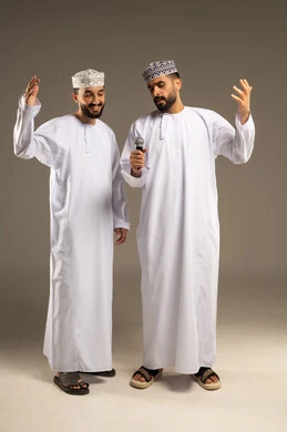 Presentation of audio performances, expressions of integration and interaction with music, a portrait of two Arab Gulf Omani men wearing the dishdasha and kumma, one of them holding a microphone, gray background.