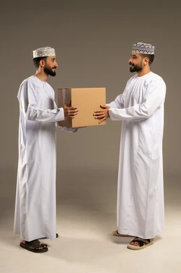 Reliable shipping and delivery process; portrait of two Omani Gulf Arab men wearing dishdashas and kumas, one of whom is handing a cardboard box to a customer, gray background