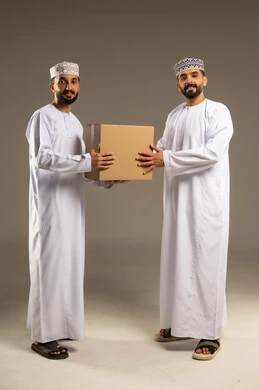 توصيل الطلبات في الوقت المحدد, بورتريه لرجلين عربيين خليجيين عمانيين يرتديان الدشداشة و الكمة يقوم أحدهما بتسليم صندوق كرتون للعميل و يقومان بالنظر الى الكاميرا بابتسامة, خلفية رمادية