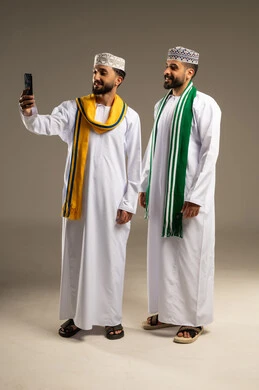 توثيق اللحظات السعيدة, ارتداء الأوشحة التشجيعية, بورتريه لرجلين عربيين خليجيين عمانيين يرتديان الدشداشة و الكمة يلتقط أحدهما سيلفي عبر الهاتف المحمول, خلفية رمادية
