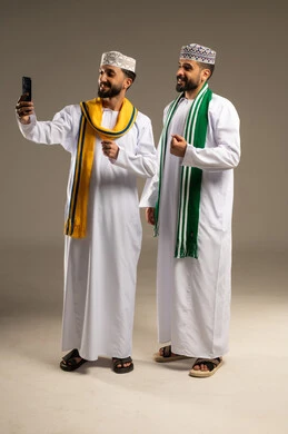 رفع قبضة اليد لتسجل هدف، التقاط صورة سيلفي, ارتداء الأوشحة التشجيعية, بورتريه لرجلين عربيين خليجيين عمانيين يرتديان الدشداشة و الكمة يستخدم أحدهما الهاتف المحمول, خلفية رمادية