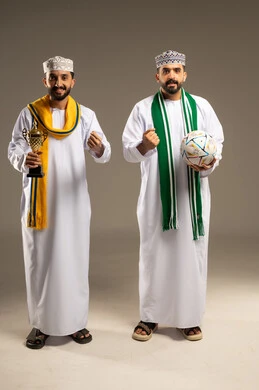 التركيز و الترقب مع المبارة, ارتداء الأوشحة التشجيعية, بورتريه لرجلين عربيين خليجيين عمانيين يرتديان الدشداشة و الكمة يمسك أحدهما بالكرة و الاخر يحمل كأس ذهبي و يرفعان قبضتيهما علامة النصر, خلفية رمادية