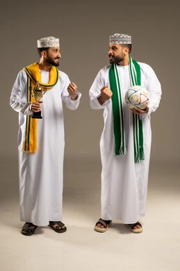 Looking with gestures of challenge, wearing supportive scarves, a portrait of two Arab Gulf Omani men wearing the dishdasha and kumma, one holding a ball and the other holding a golden cup, raising their fists as a sign of victory, gray background.