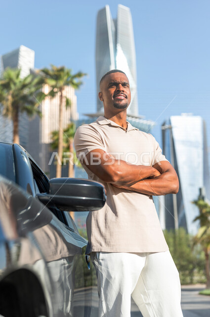 شاب ذو بشرة سمراء عربي خليجي سعودي يقف بجانب سيارته في أحد شوارع الرياض ، الاستمتاع و الإنبهار بجمال المدينة ، قضاء وقت ممتع في مدينة الملك عبد الله المالية ( كافد ) في الرياض في المملكة العربية السعودية