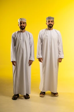 النظر الى الكاميرا بابتسامه, عرض ازياء رجاليه عمانيه شعبيه, الأناقة و الاهتمام بالمظهر الخارجي، بورتريه لرجلين عربيين خليجيين عمانيين يرتديان الدشداشة و الكمة و تظهر عليهم ايماءات الثقة, خلفية صفراء