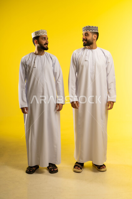 الاستجمام و التنزه مع الاصدقاء, الوقوف جانبا الى جنب, الاهتمام بالمظهر الخارجي، بورتريه لرجلين عربيين خليجيين عمانيين يرتديان الدشداشة و الكمة ينظران الى بعضهما بسعادة, خلفية صفراء