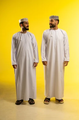 الاستجمام و التنزه مع الاصدقاء, الوقوف جانبا الى جنب, الاهتمام بالمظهر الخارجي، بورتريه لرجلين عربيين خليجيين عمانيين يرتديان الدشداشة و الكمة ينظران الى بعضهما بسعادة, خلفية صفراء