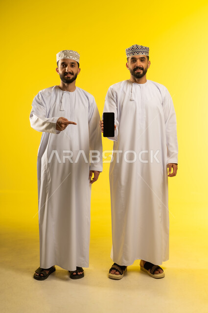 الاشارة باصبع السبابة الى الموبايل, بورتريه لرجلين عربيين خليجيين عمانيين يرتديان الدشداشة و الكمة يقوم أحدهما بعرض شاشة جوال سوداء، دمج التقنيات التكنولوجية في الحياة اليومية، خلفية صفراء