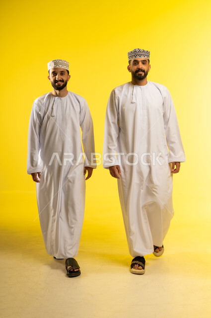 Walking confidently through the streets of Oman, enjoying time with friends, a portrait of two Omani Gulf Arab men wearing dishdashas and kumas, looking happily at the camera, yellow background