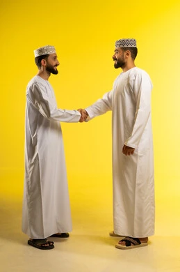 Warm greeting and handshake, a successful social relationship, exchanging greetings and peace, a portrait of two Arab Gulf Omani men wearing the dishdasha and kumma, greeting each other, gestures of happiness and joy, yellow background.