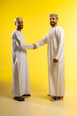 العلاقات الاجتماعية الناجحة، المصافحة و الترحيب و الاستقبال, إلقاء التحية و السلام, بورتريه لرجلين عربيين خليجيين عمانيين يرتديان الدشداشة و الكمة و يسلمان على بعضهما، ايماءات السعادة و السرور, خلفية صفراء