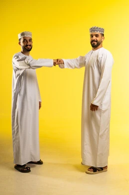 النظر الى الكاميرا ببشاشة, الصداقه و تكوين علاقات, بورتريه لرجلين عربيين خليجيين عمانيين يرتديان الدشداشة و الكمة و يقومان بتلامس قبضتي اليدين, ايماءات السعادة و السرور, خلفية صفراء