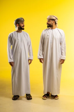 الوقوف جانبا الى جنب, الأناقة و الاهتمام بالمظهر الخارجي، بورتريه لرجلين عربيين خليجيين عمانيين يرتديان الدشداشة و العمامة ينظران الى بعضهما,  ايماءات السعادة و السرور, خلفية صفراء