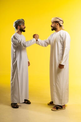 Handshaking, welcoming, and receiving, greeting and peace, friendship and building relationships, a portrait of two Arab Gulf Omani men wearing the dishdasha and turban, greeting each other, gestures of happiness and joy, yellow background.