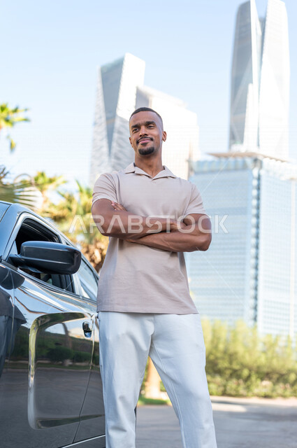 A dark-skinned young Arab Gulf Arab man stands next to his car in a street in Riyadh, enjoying and fascinated by the beauty of the city, spending a good time in King Abdullah Financial City (KAFD) in Riyadh in the Kingdom of Saudi Arabia