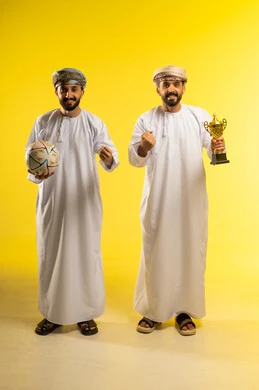 ايماءات السعادة بالانتصار, أندية رياضية مختلفة، مشاهدة و متابعة مباراة كرة القدم، بورتريه لرجلين عربيين خليجيين عمانيين يرتديان الدشداشة و العمامة و يمسك أحدهما بالكرة و الاخر يحمل كأس ذهبي, خلفية صفراء