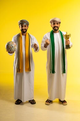 أنديه رياضية مختلفة، الشغف و تشجيع الفريق، مشاهدة و متابعة مباراة كرة القدم، بورتريه لرجلين عربيين خليجيين عمانيين يرتديان الدشداشة و العمامة و يمسك أحدهما بالكرة و الاخر يحمل كأس ذهبي, ارتداء الأوشحة التشجيعية, خلفية صفراء