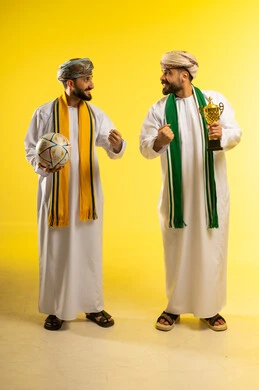 Wearing supportive scarves, different sports clubs, passion and cheering for the team, watching and following a football match, a portrait of two Arab Gulf Omani men wearing the dishdasha and turban, one holding a ball and the other holding a golden cup, yellow background.