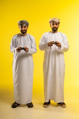أنشطة ترفيهية ممتعة, بورتريه لرجلين عربيين خليجيين عمانيين يرتديان الدشداشة و العمامة يمسكان عصا التحكم, المتعة و التسلية بألعاب الفيديو برفقة الأصدقاء, إيماءات وجه ويدين تدل على الانتصار، خلفية صفراء