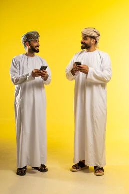 تصفح التطبيقات الذكية، التواصل عبر الإنترنت، بورتريه لرجلين عربيين خليجيين عمانيين يرتديان الدشداشة و العمامة يستخدمان الموبايل, الإستمتاع مع الأصدقاء, ايماءات السعادة و السرور, خلفية صفراء