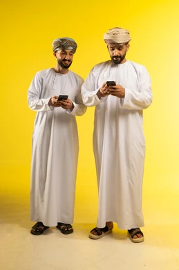 استخدام جهاز تقني حديث، قضاء اوقات ممتعة بتبادل الاخبار، الإجابة والرد على الاتصالات، بورتريه لرجلين عربيين خليجيين عمانيين يرتديان الدشداشة و العمامة يستخدمان الموبايل, الإستمتاع مع الأصدقاء, ايماءات السعادة و السرور, خلفية صفراء