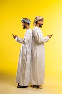 Following entertainment programs, integrating technology into daily life, a portrait of two smiling Arab Gulf Omani men wearing dishdasha and turban standing back to back using a mobile phone, making a video call with family, having a good time, yellow background.