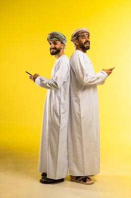 الوقوف ظهرا لظهر, دمج التكنولوجيا بالحياة اليومية, بورتريه لرجلين عربيين خليجيين عمانيين مبتسمين يرتديان الدشداشة و العمامة و يستخدمان الهاتف المحمول, إجراء مكالمة فيديو مع الأهل، قضاء وقت ممتع، خلفية صفراء