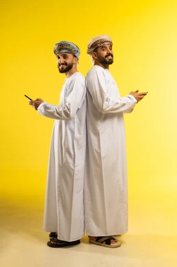 Integrating technology into daily life, a portrait of two smiling Arab Gulf Omani men wearing dishdasha and turban standing back to back using a mobile phone, having a good time, with a yellow background.