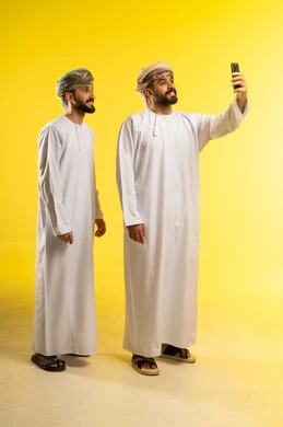 التقاط صور سيلفي، إجراء مكالمة فيديو مع الأهل، الاستمتاع بمتابعة البرامج الترفيهية، قضاء وقت ممتع، بورتريه لرجلين عربيين خليجيين عمانيين يرتديان الدشداشة و العمامة يستخدم أحدهما الموبايل لأخذ صورة تذكارية, خلفية صفراء