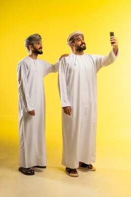 إلتقاط صور سيلفي تذكارية، قضاء وقت ممتع، الاستمتاع بمتابعة البرامج الترفيهية،  بورتريه لرجلين عربيين خليجيين عمانيين يرتديان الدشداشة و العمامة يستخدم أحدهما الموبايل لأخذ صورة, خلفية صفراء