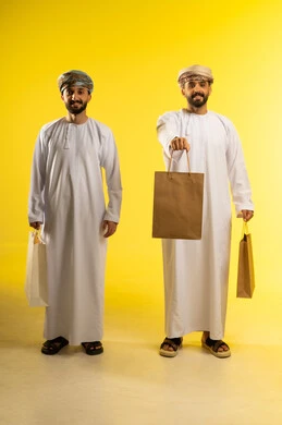 Season of promotions, discounts, and clearance sales, commercial purchases and gifts, the joy of shopping, a portrait of two Arab Gulf Omani men wearing the dishdasha and turban carrying paper bags, yellow background.