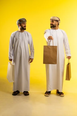 Commercial purchases and gifts, the joy of shopping, benefiting from the season of offers, discounts, and clearance sales, a portrait of two Arab Gulf men from Oman wearing the dishdasha and turban carrying paper bags, yellow background.