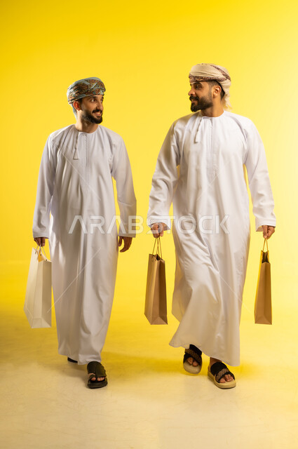 The enjoyment of shopping, benefiting from the season of offers and sales, commercial purchases and gifts, shopping and taking necessary supplies, a portrait of two Arab Gulf Omani men wearing the dishdasha and turban carrying paper bags, yellow background.