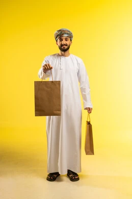 Taking advantage of seasonal offers and discounts, buying supplies and shopping at commercial malls, a portrait of an Arab Gulf Omani man wearing a dishdasha and a turban holding paper bags in his hand and looking at the camera, with a yellow background.