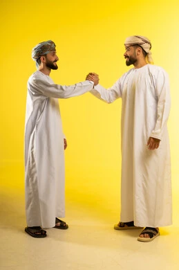 مفهوم الاستقبال و الترحيب, إلقاء التحيه و السلام, الصداقه و تكوين علاقات, بورتريه لرجلين عربيين خليجيين عمانيين يرتديان الدشداشة و العمامة يقومان بمصافحة بعضهما، ايماءات السعادة و السرور, خلفية صفراء