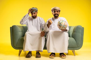 الاستمتاع بمشاهدة الفريق المفضل، بورتريه لرجلين عربيين خليجيين عمانيين يرتديان الدشداشة و العمامة يجلسان على أريكة مريحة يمسك أحدهما كرة بيده و تظهر عليه ايماءات السعادة بالفوز و يضع الاخر يده على رأسه بايماءات و الخسارة, خلفية صفراء