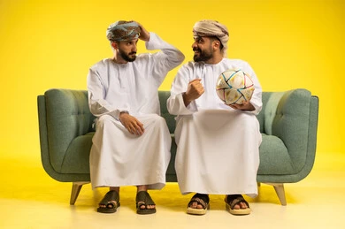 Matches and sports, a portrait of two Arab Gulf Omani men wearing the dishdasha and turban sitting on a comfortable sofa, one holding a ball in his hand showing gestures of happiness from winning, while the other has his hand on his head showing gestures of loss, with a yellow background.