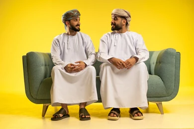 جلسة وناسة و تعارف، مناقشة الأخبار و التحدث مع الأصدقاء، بورتريه لرجلين عربيين خليجيين عمانيين يرتديان الدشداشة و العمامة يجلسان على أريكة مريحة  و يتبادلان أطراف الحديث, إيماءات تدل على السعادة, خلفية صفراء