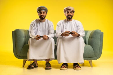 المشاركة بالألعاب التقنية المتطورة, بورتريه لرجلين عربيين خليجيين عمانيين يرتديان  الدشداشة و العمامة يمسكان عصا التحكم, الاستمتاع بالفيديو جيمز برفقة الأصدقاء,  إيماءات وجه تدل على الإندماج مع اللعبة,  خلفية صفراء