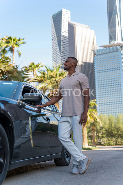 شاب ذو بشرة سمراء عربي خليجي سعودي يقف بجانب سيارته في أحد شوارع الرياض ، الاستمتاع و الإنبهار بجمال المدينة ، قضاء وقت ممتع في مدينة الملك عبد الله المالية ( كافد ) في الرياض في المملكة العربية السعودية