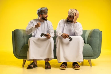 تعابير وجه و يد تدل على الخسارة و الحزن، الاندماج و التفاعل بلعب ألعاب الفيديو، بورتريه لرجلين عربيين خليجيين عمانيين يرتديان الدشداشة و العمامة يمسكان عصا التحكم, العالم الرقمي الترفيهي, مفهوم الفوز و السعادة, خلفية صفراء