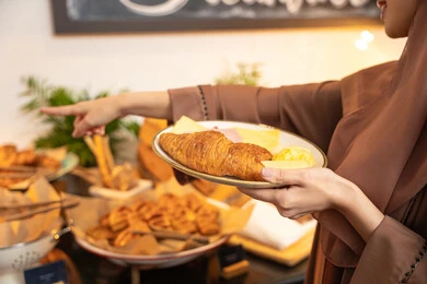 Joy and enjoyment outside the home, delicious and fresh foods and pastries, a variety of different dishes, choosing a breakfast dish from the open buffet, spending enjoyable times on the weekend, a close-up shot of an Emirati Gulf Arab woman holding a plate of food. Joy and enjoyment outside the home, delicious and fresh foods and pastries, a variety of different dishes, choosing a breakfast dish from the open buffet, spending enjoyable times on the weekend, a close-up shot of an Emirati Gulf Arab woman holding a plate of food.