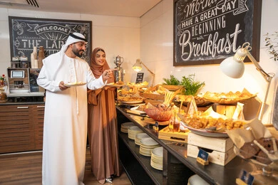 Choosing a breakfast dish, enjoying breakfast, spending enjoyable times on the weekend, the concept of a happy family, an Emirati Arab couple wearing traditional Emirati attire serving food from the open buffet in a luxury restaurant, a variety of different dishes of delicious and fresh foods and pastries.