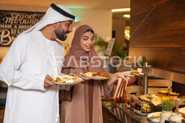 اختيار طبق الفطور ، الاستمتاع بتناول طعام الفطور، قضاء اوقات ممتعه في عطلة نهاية الاسبوع,مفهوم الاسره السعيده, زوجان عربيان اماراتيان  يرتديان الزي التقليدي الاماراتي يغرفان الطعام من البوفيه المفتوح في مطعم فاخر, اطباق متنوعه ومختلفه من المأكولات والمخبوزات الشهية والطارجة