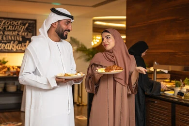 السعادة والاستمتاع خارج المنزل، المأكولات والمخبوزات الشهية والطازجة ،اطباق متنوعه ومختلفه ،اختيار طبق الفطور من البوفيه المفتوح، قضاء اوقات ممتعه في عطلة نهاية الاسبوع, زوجان عربيان اماراتيان  يرتديان الزي التقليدي الاماراتي يحملان اطباق الطعام